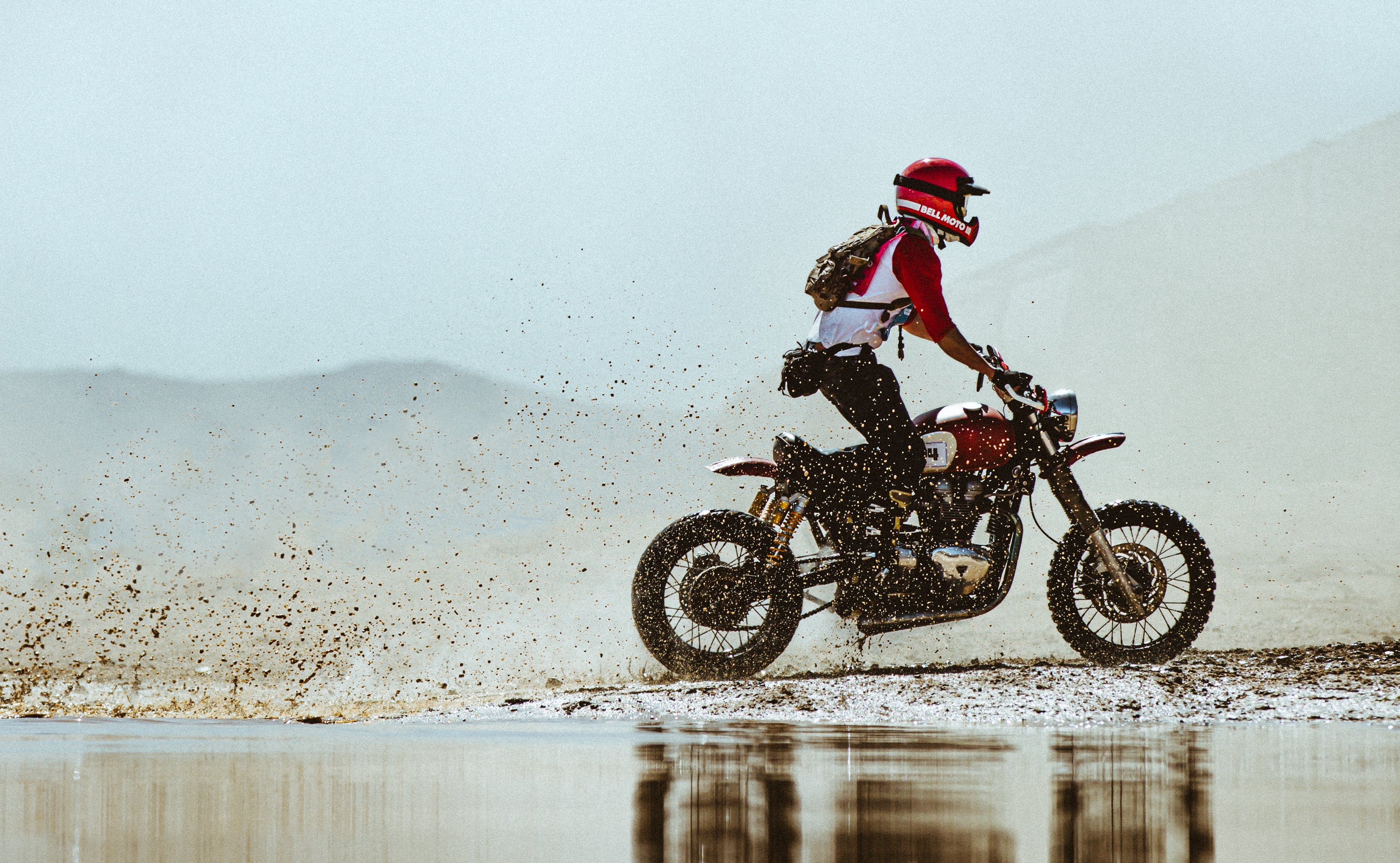 A side view of a rider on a red and black off-road motorcycle, riding on a sandy beach with water reflection in the foreground.